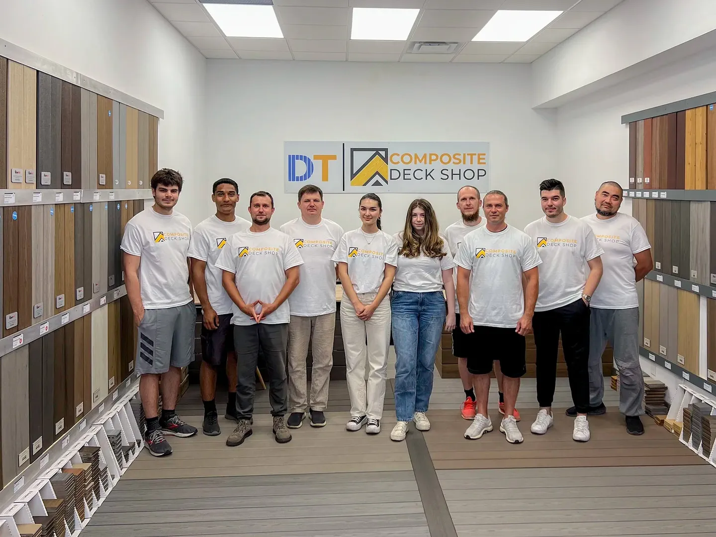 Ten members of the Composite Deck Shop team in matching T-shirts, standing between walls of decking samples under a DT and Composite Deck Shop sign in the Vaughan showroom
