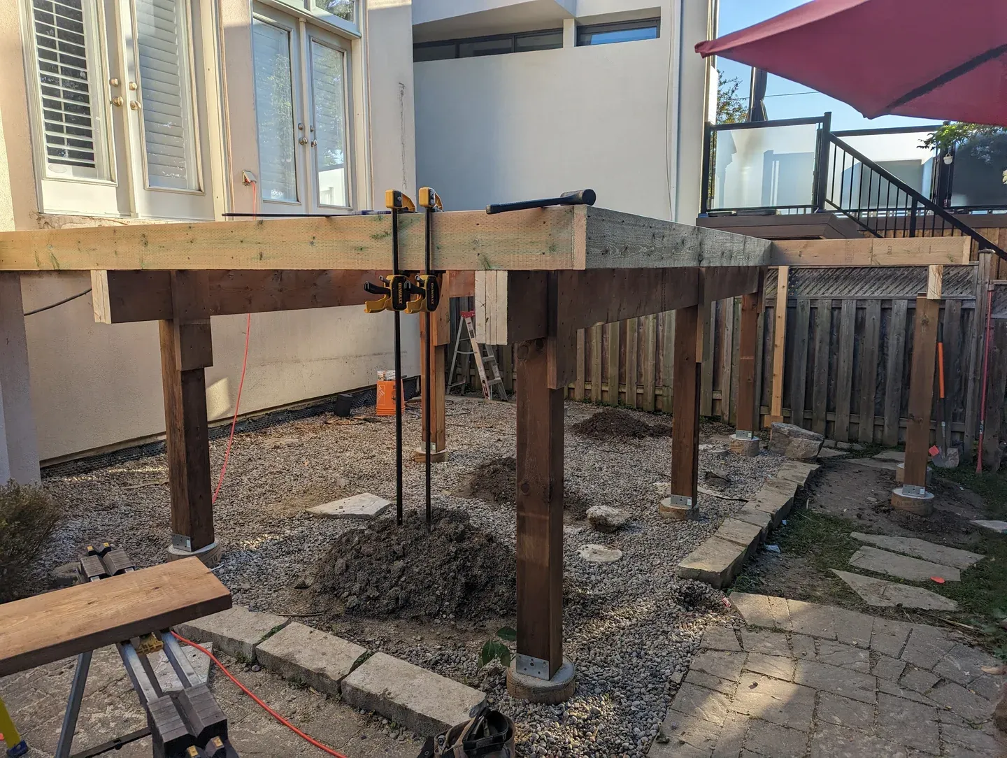 Deck frame under construction beside a house, with timber posts on concrete piers and perimeter framing clamped in place