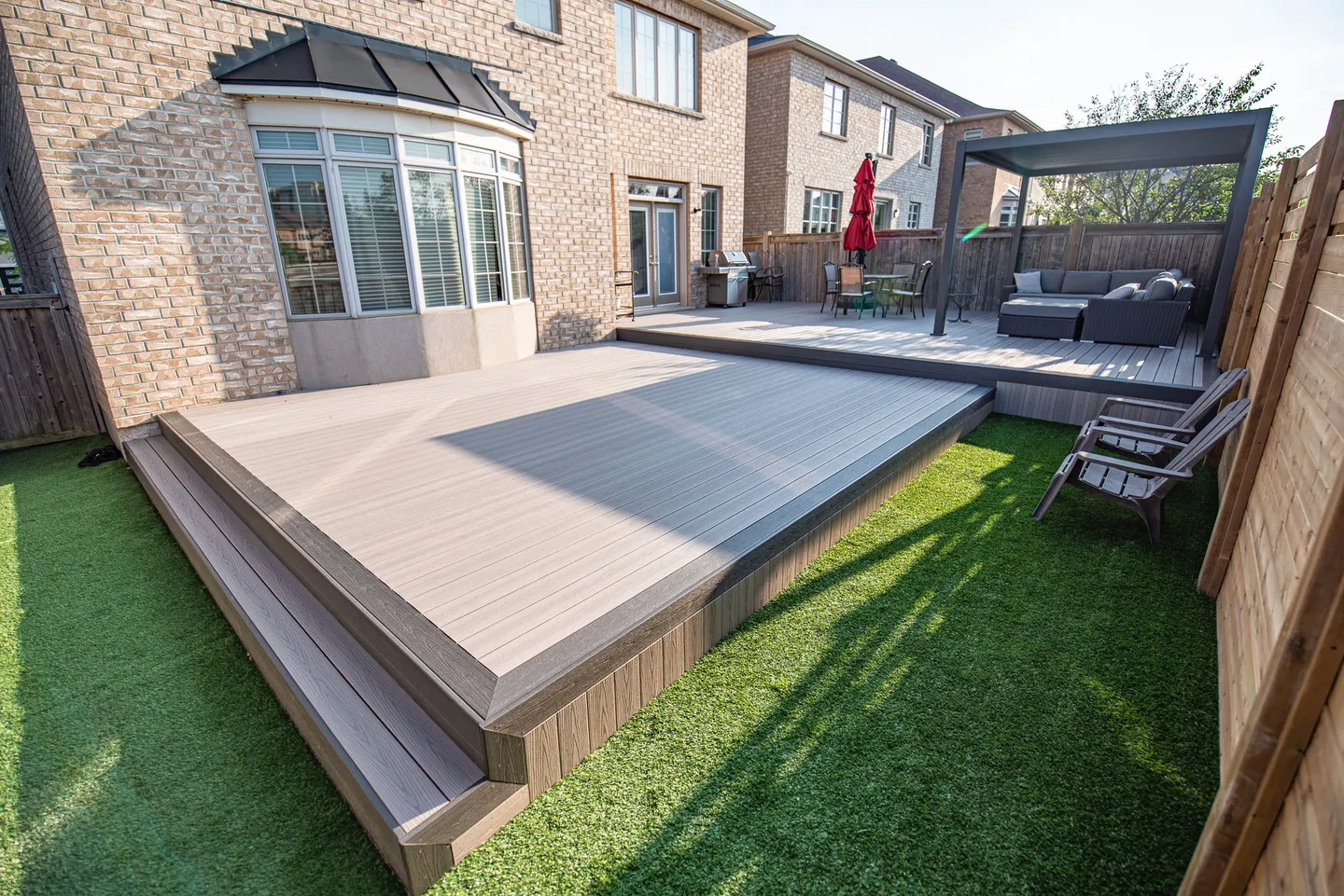 Grey Fiberon deck with a contrasting perimeter, broad steps, a pergola and adjoining artificial turf.