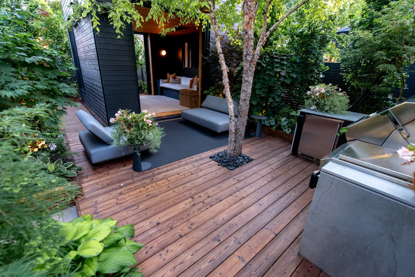Wood deck arranged around a tree and planted borders, with lounge seating and an open garden room.