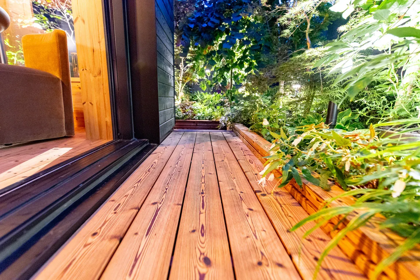 Close view of knotted wood decking alongside an open garden-room doorway and leafy planting.