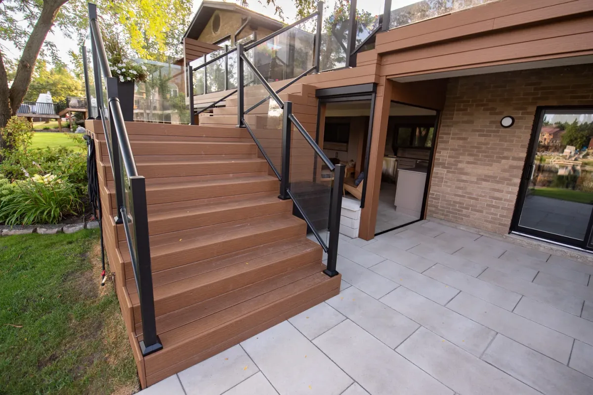 Brown deck stairs with framed tread edges, glass guards and dark posts beside a stone patio